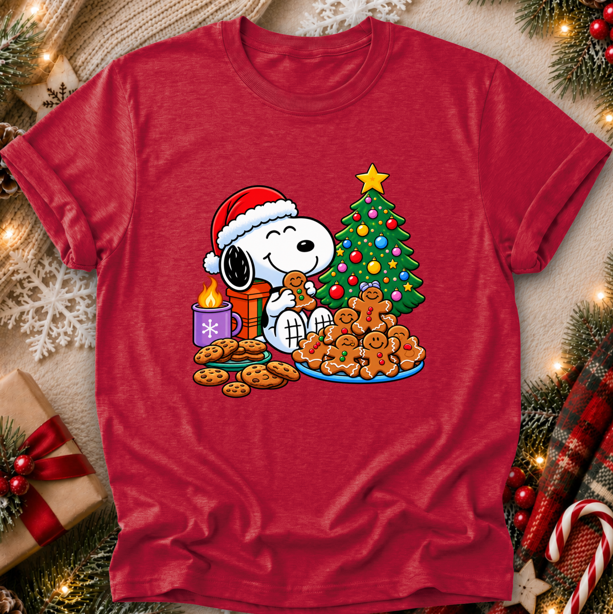 A red short-sleeve crewneck t-shirt featuring a festive graphic of Snoopy wearing a Santa hat, eating a cookie next to a decorated Christmas tree, cookies, and a gift box. The shirt is displayed in a flat-lay holiday setting with pine branches, lights, and candy canes.