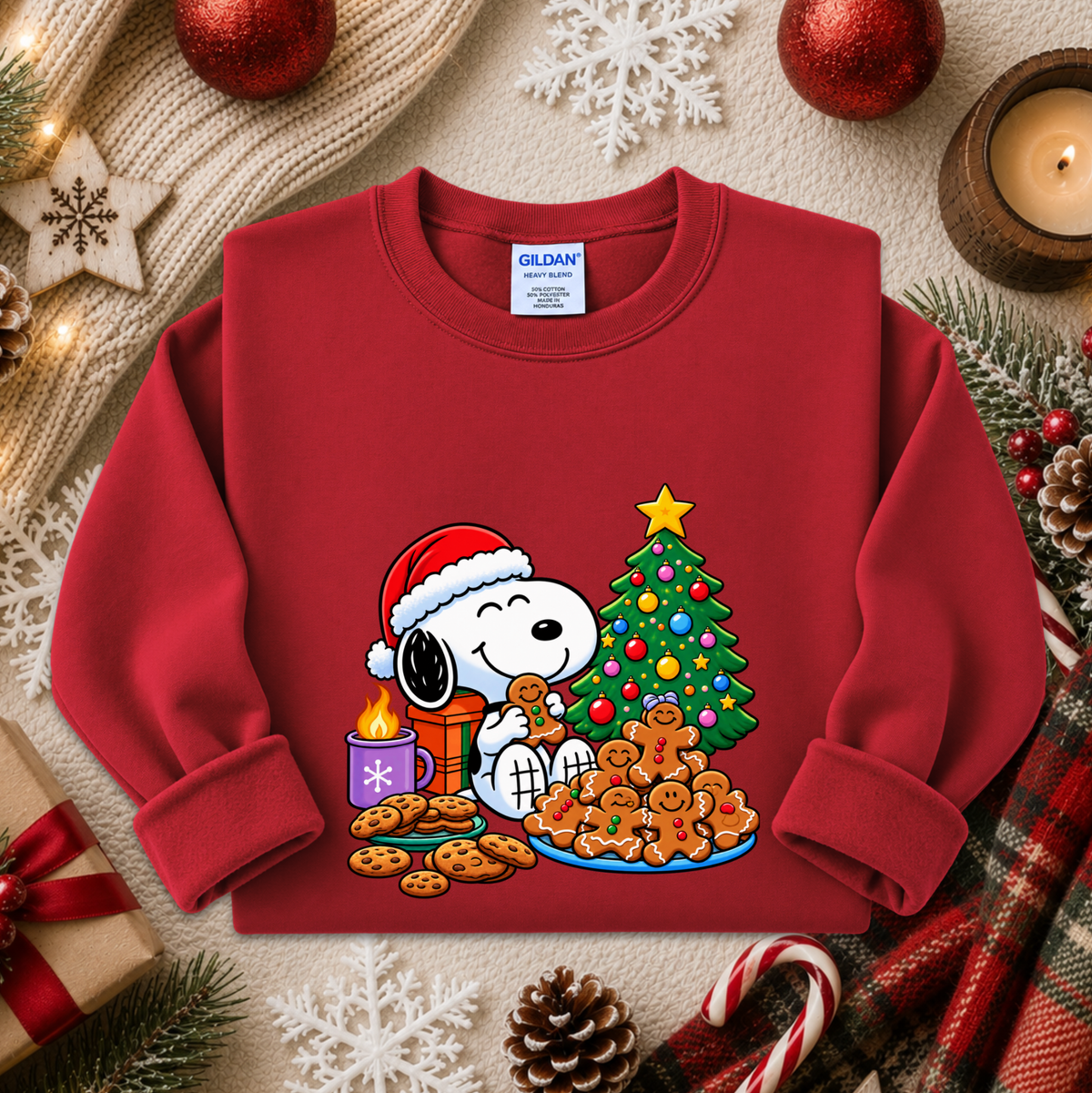 A red long-sleeve crewneck sweatshirt featuring a festive Christmas graphic of Snoopy and Woodstock with cookies, a decorated tree, and a Santa hat, displayed in a cozy holiday flat-lay setting.