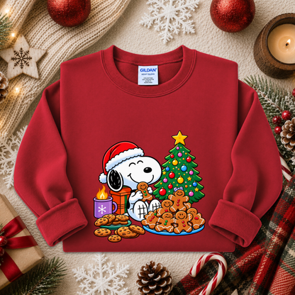 A red long-sleeve crewneck sweatshirt featuring a festive Christmas graphic of Snoopy and Woodstock with cookies, a decorated tree, and a Santa hat, displayed in a cozy holiday flat-lay setting.