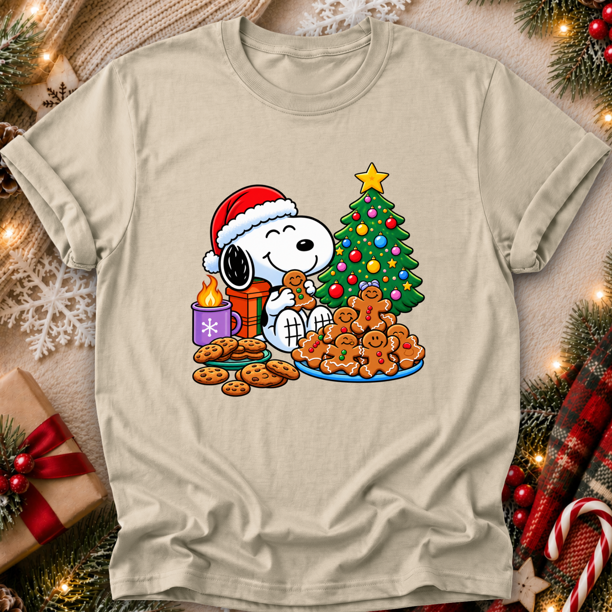 A beige crewneck t-shirt featuring a colorful graphic of Snoopy and Woodstock celebrating Christmas with cookies, a tree, and a gift box, displayed on a cozy holiday-themed background.