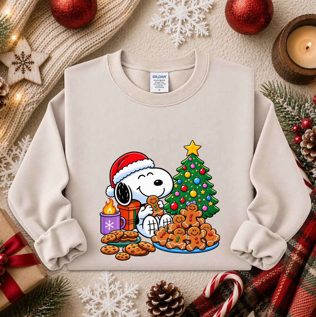 A light beige crewneck sweatshirt featuring a colorful graphic of Snoopy and Woodstock by Christmas, styled in a cozy holiday flat-lay with a knitted sweater, candle, gingerbread cookies, and pinecones.