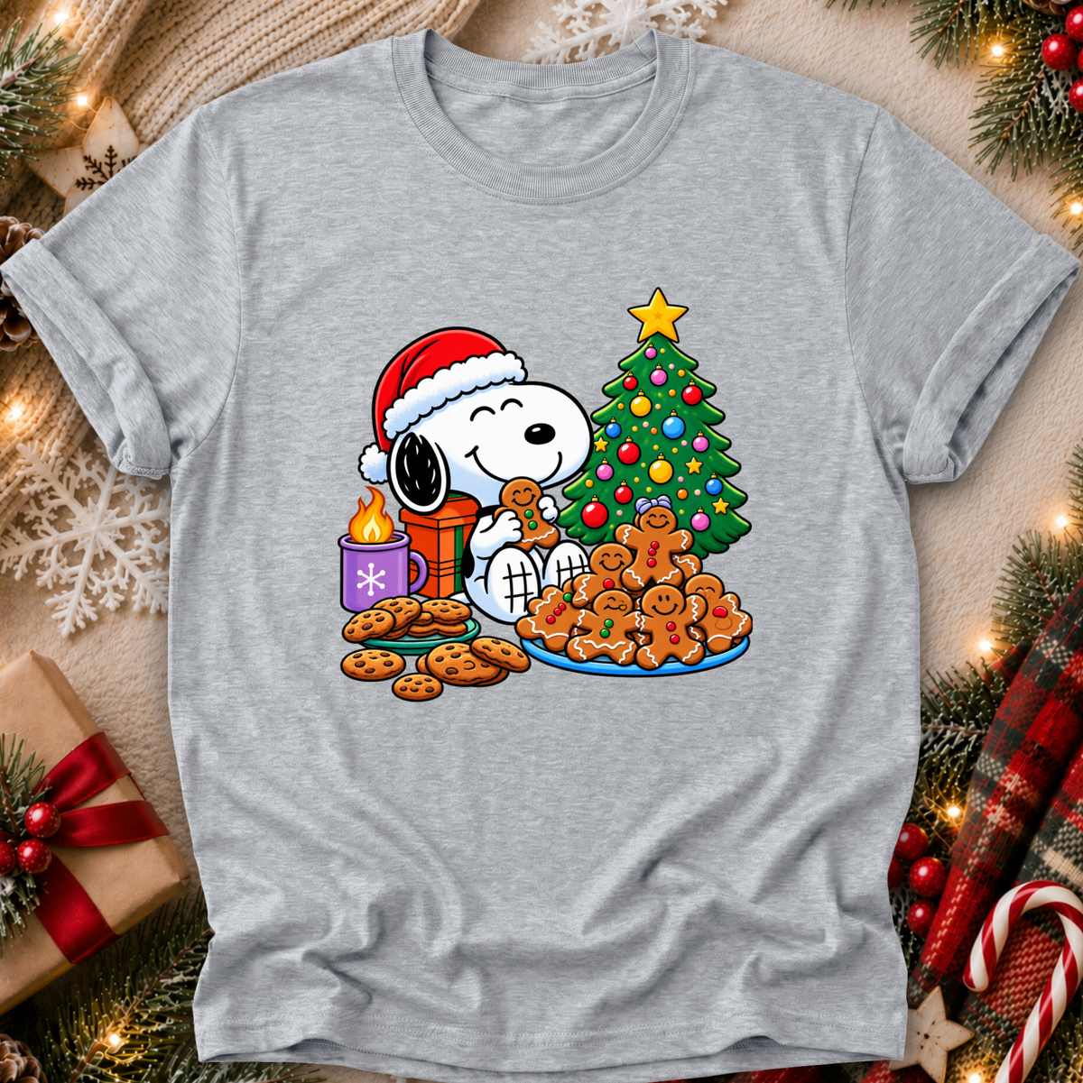 A heather gray short-sleeve t-shirt featuring a colorful graphic of Snoopy and Woodstock celebrating Christmas with cookies, a decorated tree, and a candle. The shirt is displayed in a flat-lay setting surrounded by festive holiday decorations like pine branches, lights, and a wrapped gift box.