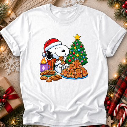 A white short-sleeve crewneck t-shirt featuring a colorful Christmas graphic of Snoopy wearing a Santa hat, eating gingerbread cookies next to a decorated tree and a gift box, styled in a cozy holiday flat-lay setting.