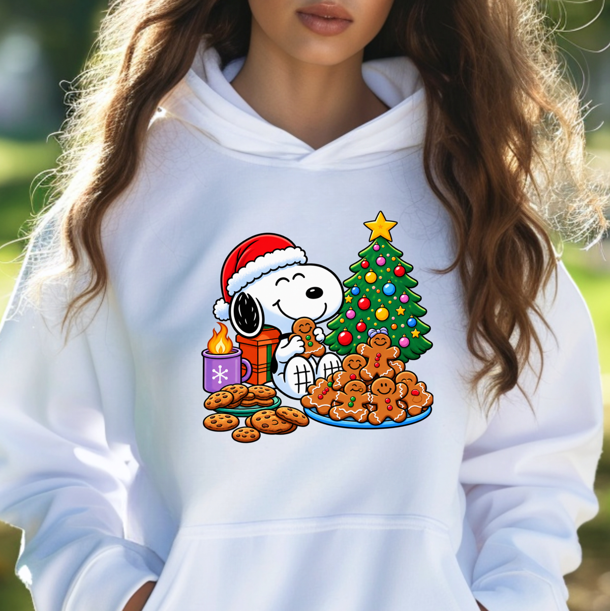 A person wearing a white pullover hoodie with a colorful Christmas graphic of Snoopy and Woodstock by a decorated tree, cookies, and gingerbread men, photographed outdoors with a blurred green background.