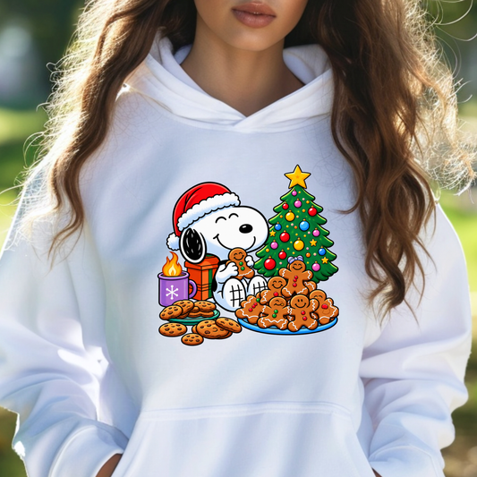 A person wearing a white pullover hoodie with a colorful Christmas graphic of Snoopy and Woodstock by a decorated tree, cookies, and gingerbread men, photographed outdoors with a blurred green background.