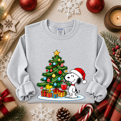 A light gray long-sleeve sweatshirt featuring a festive Christmas graphic of Snoopy and Woodstock under a decorated tree with presents. The sweatshirt is styled in a cozy holiday flat-lay with red ornaments, a candle, pinecones, and a plaid scarf.