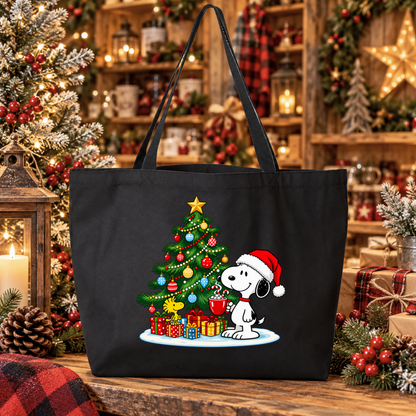 A black canvas tote bag featuring a festive Christmas graphic of Snoopy and Woodstock under a decorated tree with wrapped presents. The bag is displayed on a rustic wooden table surrounded by holiday decorations, including a lit Christmas tree, lanterns, and plaid textiles.