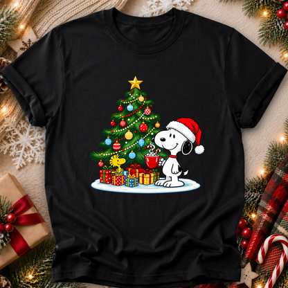 A black short-sleeve crewneck t-shirt featuring a festive Christmas graphic of Snoopy wearing a Santa hat next to a decorated Christmas tree with wrapped presents. The shirt is displayed in a styled flat-lay setting with holiday decorations including pine branches, lights, a candy cane, and a gift box.