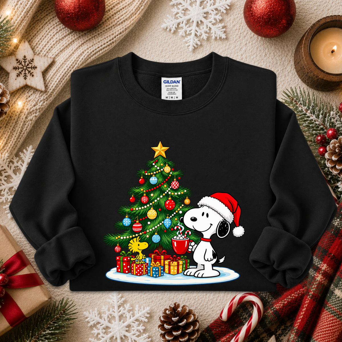 A black crewneck sweatshirt featuring a festive Christmas graphic of Snoopy wearing a Santa hat and holding a mug next to a decorated Christmas tree with presents. The sweatshirt is displayed in a styled flat-lay setting with holiday decorations including pine branches, candy canes, a candle, and a knit blanket.
