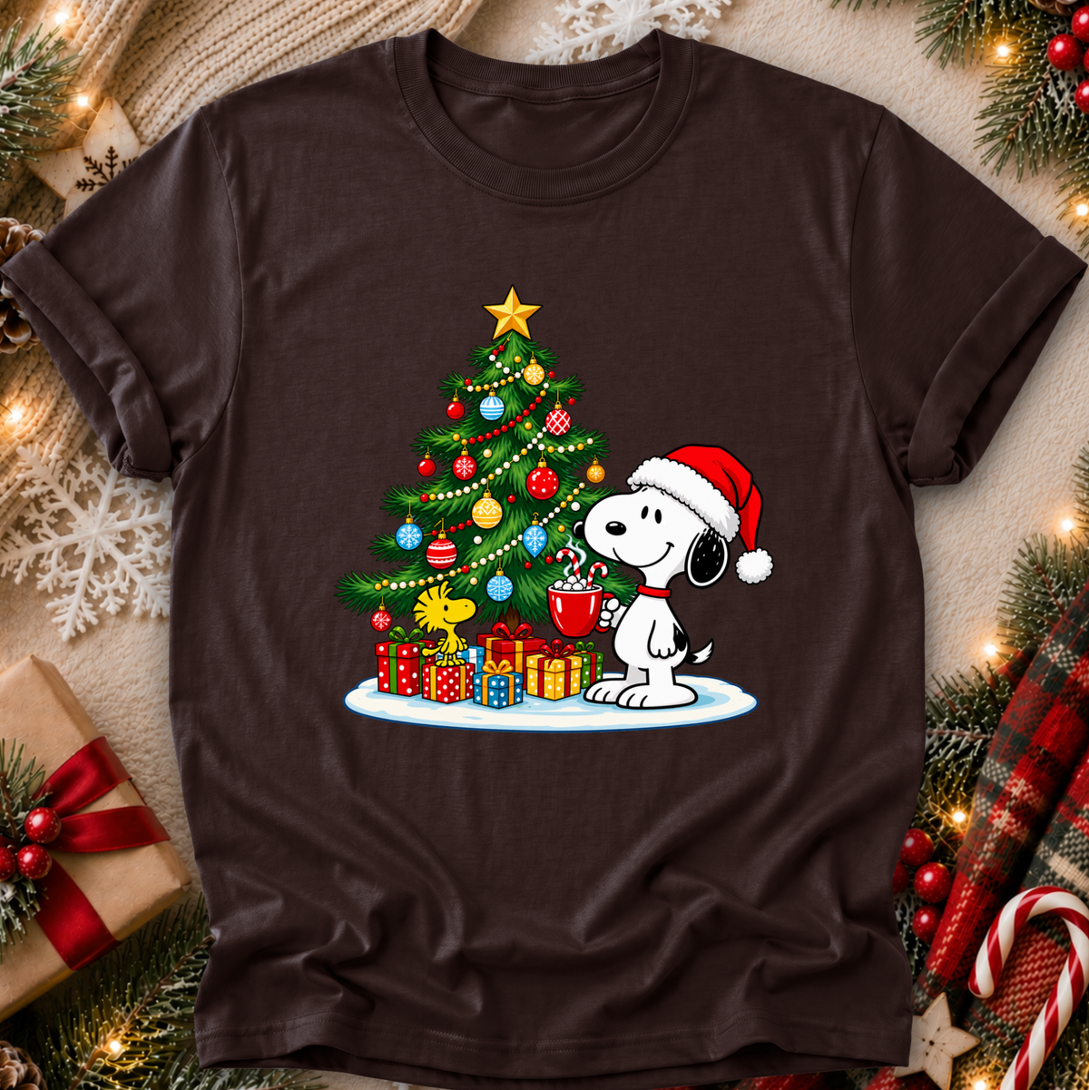 A dark brown short-sleeve crewneck t-shirt featuring a festive graphic of Snoopy wearing a Santa hat next to a decorated Christmas tree with gifts. The shirt is displayed in a flat-lay Christmas setting with pine branches, lights, and wrapped presents.