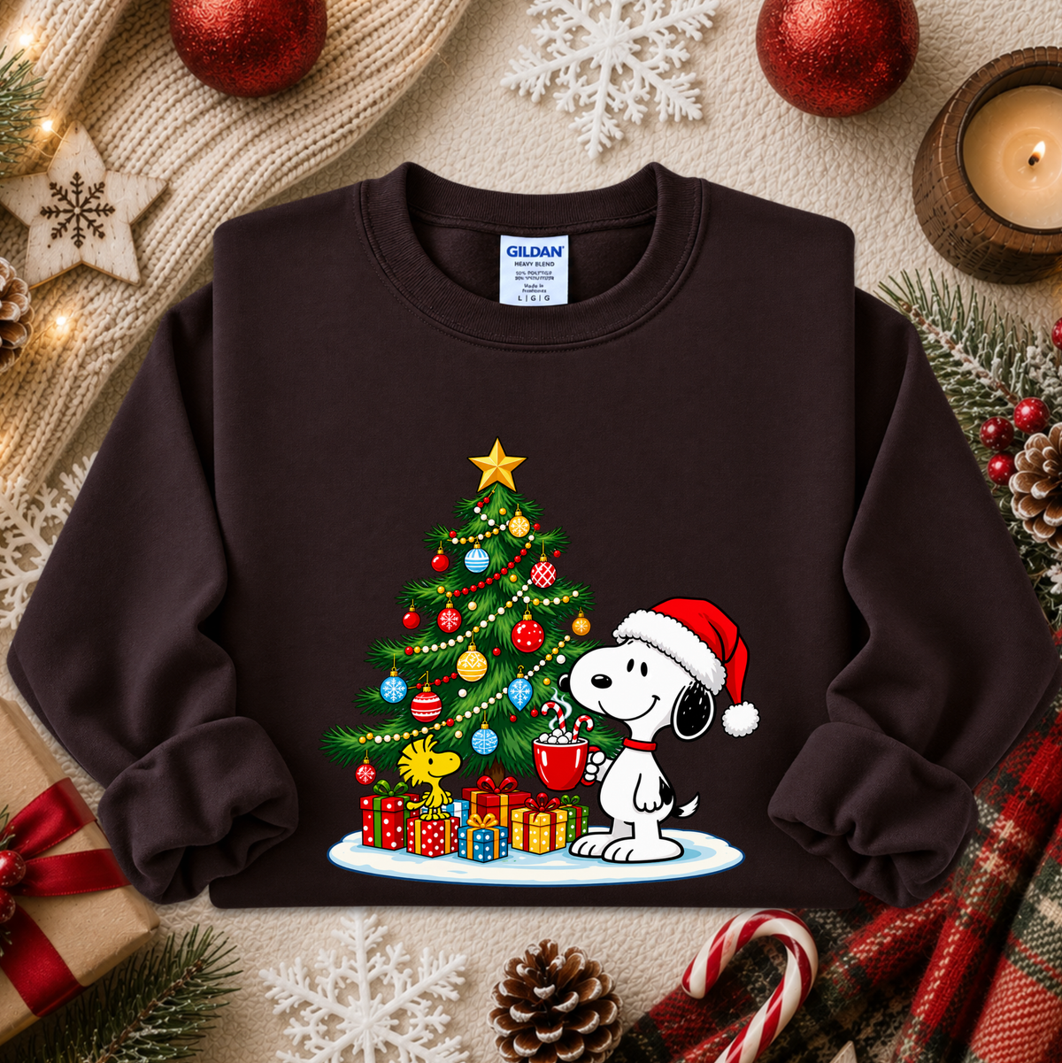 A dark brown crewneck sweatshirt featuring a festive Christmas graphic of Snoopy wearing a Santa hat next to a decorated Christmas tree with presents. The sweatshirt is styled in a cozy holiday flat-lay with a knitted sweater, candles, candy canes, pinecones, and red ornaments.