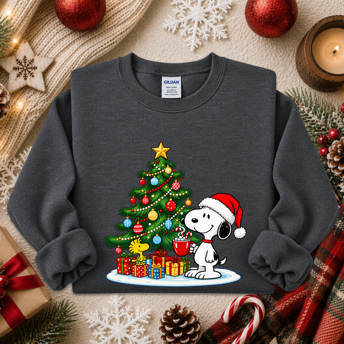 A dark gray crewneck sweatshirt featuring a festive Christmas graphic of Snoopy wearing a Santa hat next to a decorated Christmas tree with presents. The sweatshirt is styled in a cozy holiday flat-lay with pine branches, candy canes, a candle, and a knit blanket.