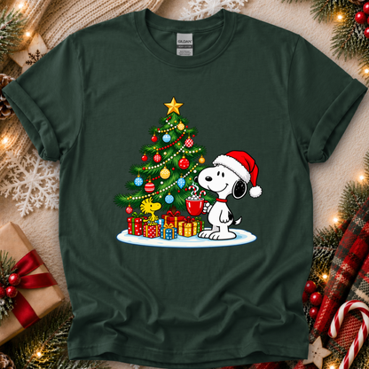A dark forest green crewneck t-shirt featuring a festive graphic of Snoopy and Woodstock by the Christmas tree, surrounded by holiday decorations like pine branches, ornaments, and wrapped gifts on a beige textured background.