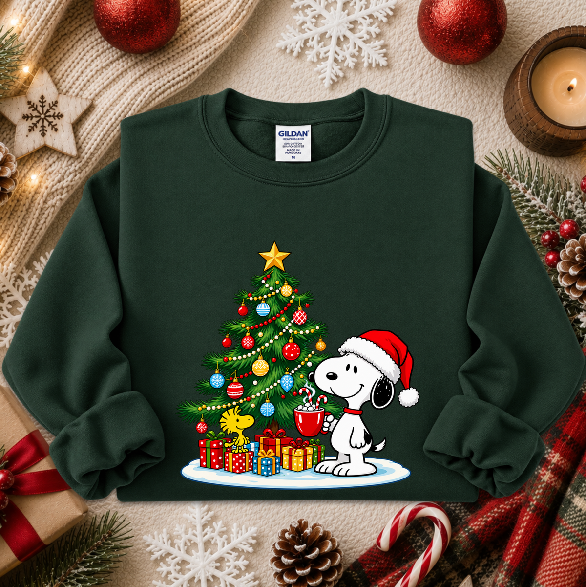 A dark green crewneck sweatshirt featuring a festive Christmas graphic of Snoopy wearing a Santa hat next to a decorated Christmas tree and wrapped presents. The sweatshirt is styled in a cozy holiday flat-lay with a knitted scarf, red ornaments, pinecones, candy canes, and a candle.