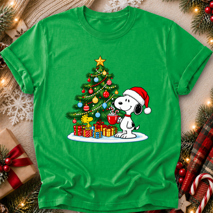 A bright green short-sleeve T-shirt laid flat with a festive Christmas graphic of Snoopy sitting by a decorated tree and holding a mug, surrounded by wrapped gifts. The shirt is styled on a cozy holiday background with lights, ornaments, pine branches, and candy canes.