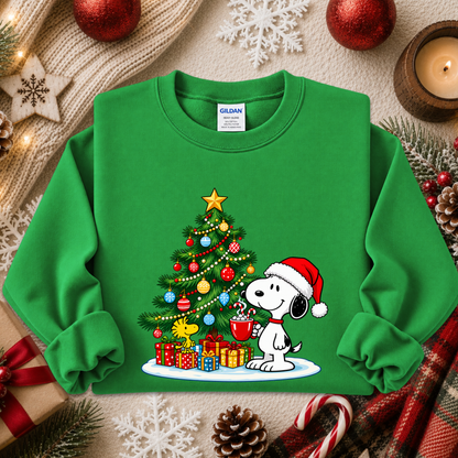 A green crewneck sweatshirt featuring a festive Christmas graphic of Snoopy wearing a Santa hat and holding a mug next to a decorated Christmas tree with presents. The sweatshirt is styled in a cozy holiday flat-lay with a knit sweater, candle, pinecone, candy cane, and red ornaments.