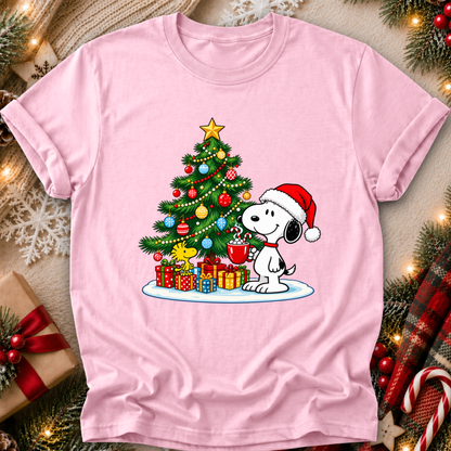 A light pink short-sleeve crewneck t-shirt featuring a festive graphic of Snoopy and Woodstock under a decorated Christmas tree with wrapped presents. The shirt is displayed in a flat-lay setting surrounded by holiday decorations like pine branches, candy canes, and a gift box.
