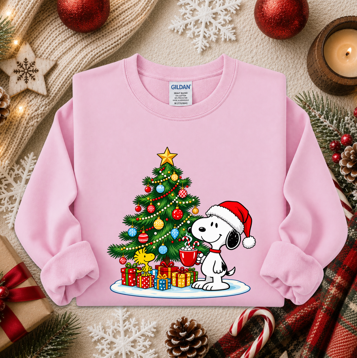 A light pink Gildan sweatshirt featuring a festive Christmas graphic of Snoopy wearing a Santa hat next to a decorated Christmas tree and presents. The sweatshirt is styled in a cozy holiday flat-lay with a knitted scarf, candles, pinecones, and candy canes.