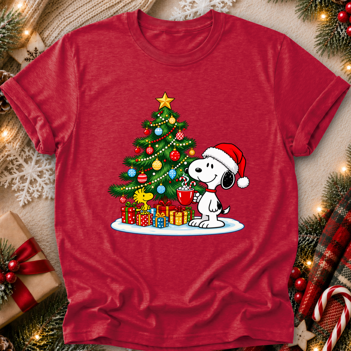 A red short-sleeve crewneck t-shirt featuring a festive Christmas graphic of Snoopy wearing a Santa hat and holding a mug next to a decorated Christmas tree with gifts. The shirt is displayed in a flat-lay setting surrounded by holiday decorations like pine branches, candy canes, and a gift box.