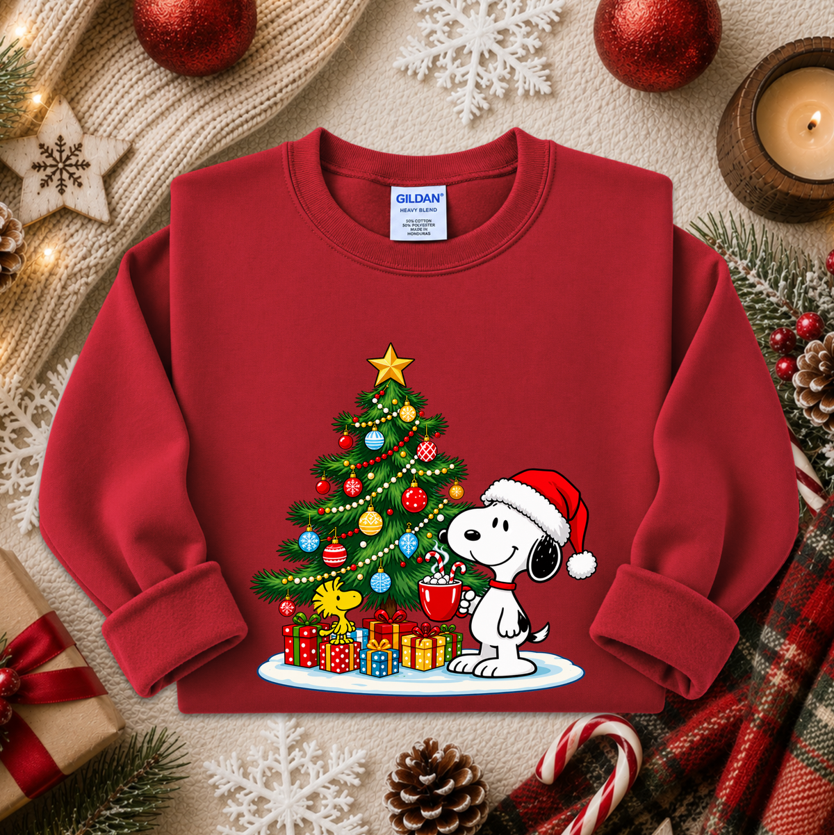A red crewneck sweatshirt featuring a festive Christmas graphic of Snoopy next to a decorated tree and wrapped gifts. The sweatshirt is displayed in a styled flat-lay arrangement with holiday props including a candle, pinecones, candy canes, and a plaid scarf.