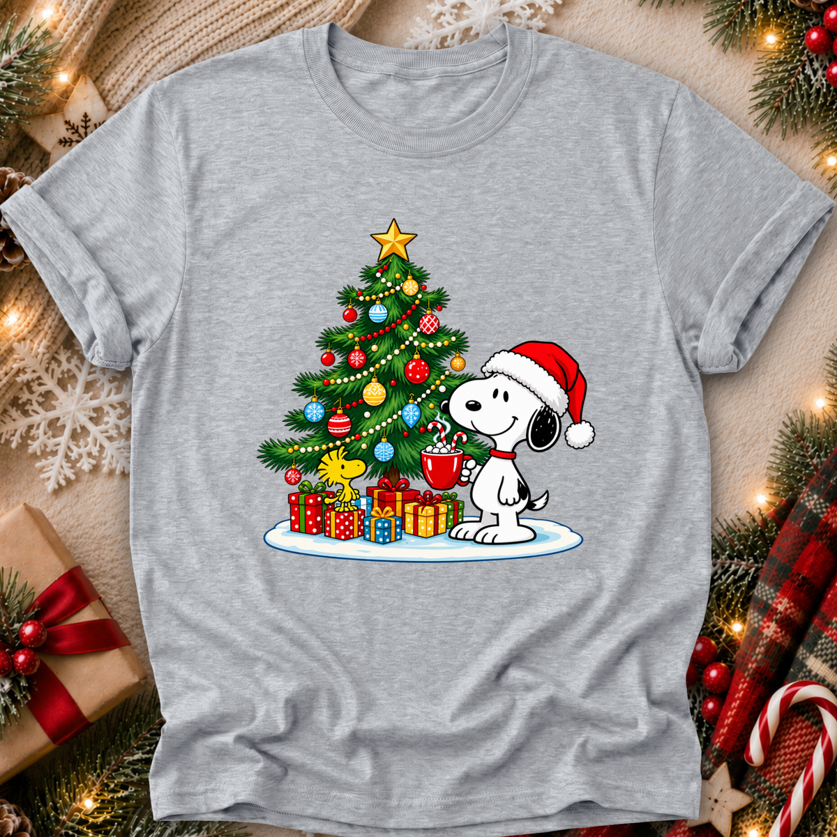 A heather gray short-sleeve T-shirt laid flat with a festive Christmas graphic of Snoopy wearing a Santa hat beside a decorated tree and wrapped presents. The shirt is styled with holiday decorations, lights, and candy canes around it.
