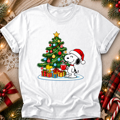 A white short-sleeve crewneck t-shirt featuring a colorful graphic of Snoopy and Woodstock under a decorated Christmas tree with wrapped gifts. The shirt is displayed in a flat-lay setting surrounded by festive holiday decorations like pine branches, candy canes, and a gift box.