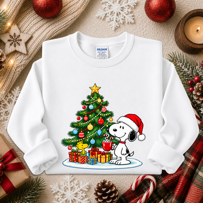 A white long-sleeve crewneck sweatshirt featuring a colorful Christmas graphic of Snoopy and Woodstock by a decorated tree with presents. The sweatshirt is styled in a cozy holiday flat-lay with red ornaments, a candle, pinecones, and a knit blanket.