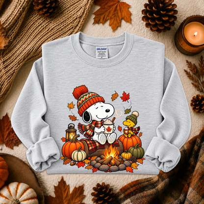 A light gray long-sleeve sweatshirt featuring a colorful autumn-themed graphic of Snoopy and Woodstock by a campfire, surrounded by pumpkins, fall leaves, pinecones, a candle, and cozy knit textiles in a styled flat-lay arrangement.