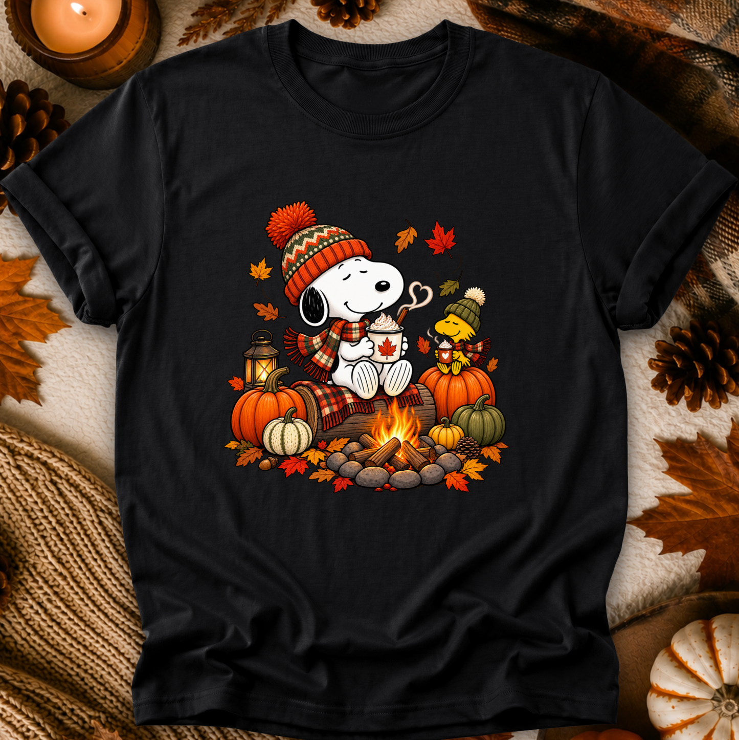 A black short-sleeve graphic t-shirt featuring Snoopy and Woodstock by a campfire in autumn colors, styled flat-lay on a cozy background with pumpkins, leaves, and a candle.