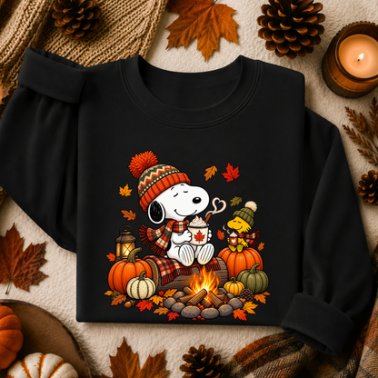 A black long-sleeve sweatshirt laid flat on a cozy autumn-themed background. The front features a colorful graphic of Snoopy and Woodstock by a campfire, wearing knit hats and scarves, with pumpkins, fall leaves, and a mug.