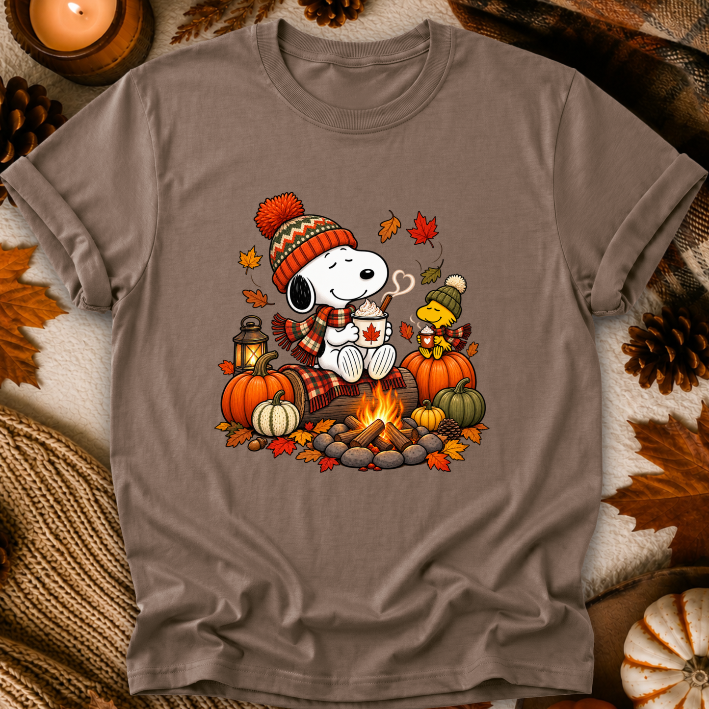 A taupe gray short-sleeve t-shirt featuring a colorful graphic of Snoopy and Woodstock in autumn clothing sitting by a campfire with pumpkins and fall leaves. The shirt is displayed in a flat-lay arrangement with cozy fall props including a candle, pinecones, and a knit blanket.