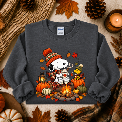 A dark gray crewneck sweatshirt featuring a colorful graphic of Snoopy and Woodstock by a campfire surrounded by pumpkins and autumn leaves. The sweatshirt is displayed in a cozy flat-lay setting with a knitted scarf, pinecones, and a lit candle.