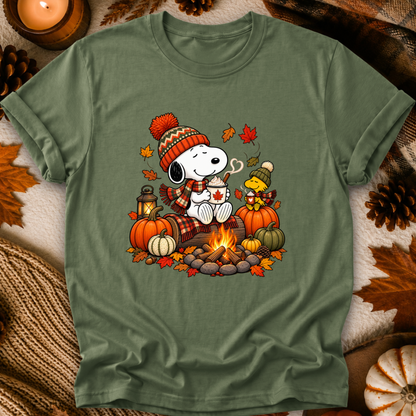 An olive green crewneck t-shirt featuring a colorful graphic of Snoopy and Woodstock sitting by a campfire surrounded by pumpkins and autumn leaves. The shirt is displayed in a flat-lay arrangement with fall-themed props like pinecones, a candle, and a knit blanket.