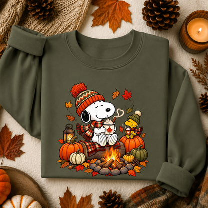 An olive green long-sleeve sweatshirt featuring a cozy autumn graphic of Snoopy and Woodstock by a campfire, surrounded by pumpkins, maple leaves, pinecones, and a candle in a styled flat-lay arrangement.