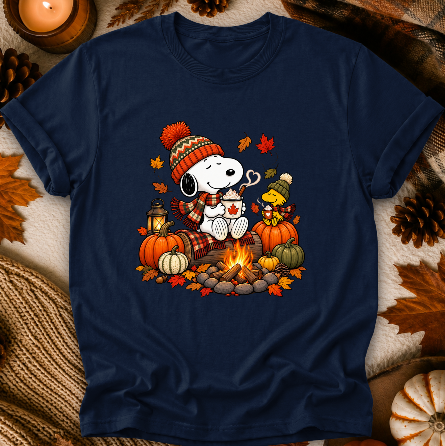 A navy blue graphic t-shirt featuring a cartoon Snoopy and Woodstock sitting by a campfire in autumn attire, surrounded by pumpkins, leaves, and pinecones, presented in a cozy flat-lay setting.