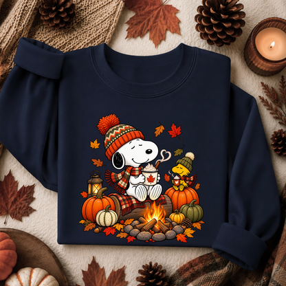 A navy blue sweatshirt featuring a colorful graphic of Snoopy and Woodstock by a campfire with pumpkins, autumn leaves, and a knit hat. The sweatshirt is styled in a flat-lay arrangement with fall-themed props including pinecones, a candle, and leaves on a neutral fabric background.