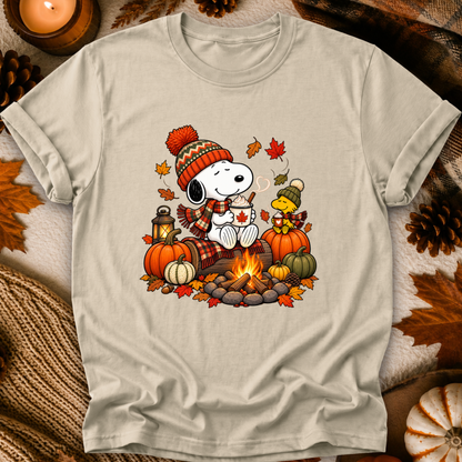 A light beige graphic t-shirt featuring a cozy fall-themed illustration of Snoopy and Woodstock by a campfire, surrounded by pumpkins, autumn leaves, and pinecones, presented in a flat-lay arrangement.