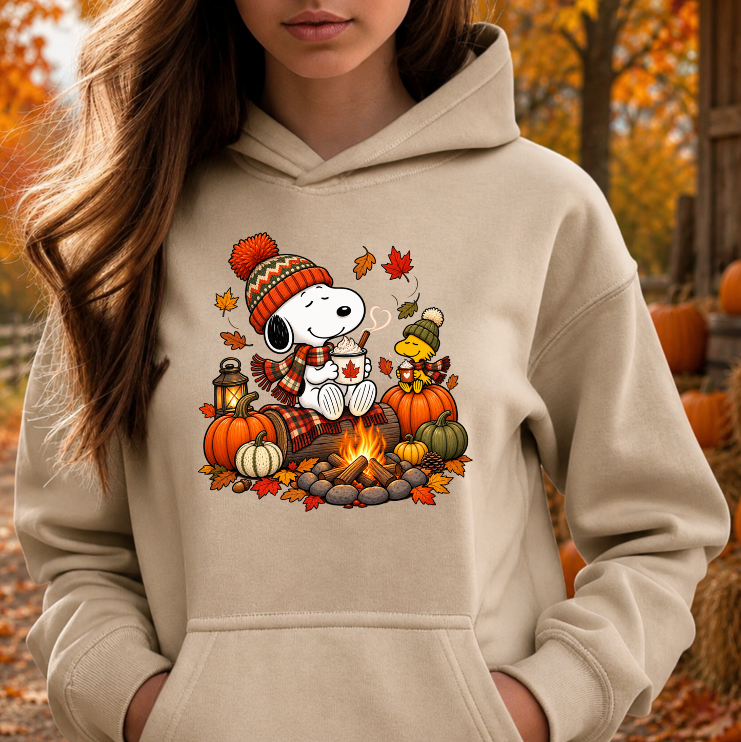 A woman wearing a beige hoodie with a colorful graphic of Snoopy and Woodstock by a campfire surrounded by pumpkins and autumn leaves. The background is a blurred outdoor autumn scene with trees and pumpkins.