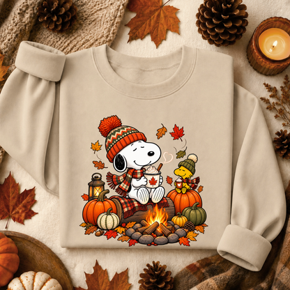 A beige long-sleeve sweatshirt laid flat with rolled cuffs, featuring a colorful autumn graphic of Snoopy and Woodstock by a campfire with pumpkins, leaves, and a lantern. The sweatshirt is styled with fall props including pinecones, autumn leaves, a candle, and a knit blanket.