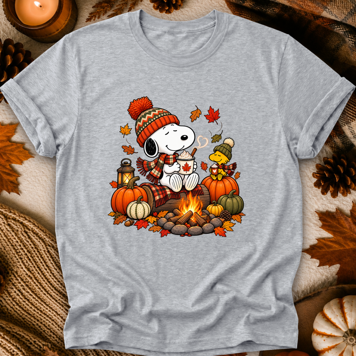 A light heather gray short-sleeve t-shirt featuring a colorful graphic of Snoopy and Woodstock by a campfire with pumpkins, autumn leaves, and a cozy fall theme, displayed on a rustic autumn-themed background.