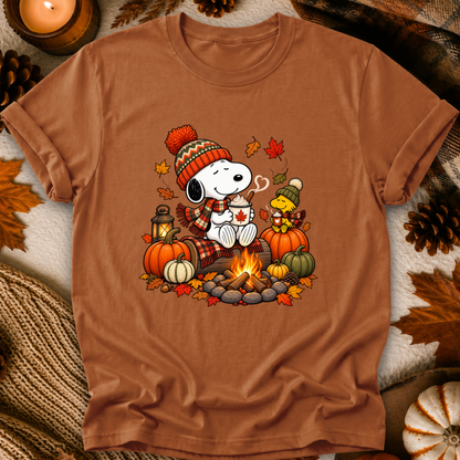 A brown short-sleeve crewneck t-shirt featuring a colorful graphic of Snoopy and Woodstock by a campfire with pumpkins, autumn leaves, and a plaid scarf, presented in a cozy flat-lay setting with pinecones and a candle.