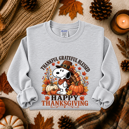 A light heather gray long-sleeve sweatshirt laid flat on a cozy autumn-themed background. The sweatshirt features a printed graphic of Snoopy wearing a pilgrim hat with pumpkins, fall leaves, and the text 'THANKFUL GRATEFUL BLESSED' and 'HAPPY THANKSGIVING.