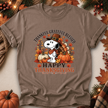 A brown short-sleeve graphic T-shirt laid flat with autumn-themed props around it. The shirt features a Snoopy illustration wearing a pilgrim hat, surrounded by pumpkins, fall leaves, and the text 'THANKFUL GRATEFUL BLESSED' and 'HAPPY THANKSGIVING'.