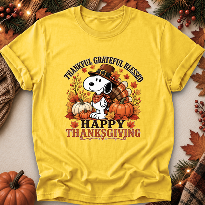 A bright yellow short-sleeve T-shirt laid flat with a Thanksgiving-themed graphic featuring Snoopy wearing a pilgrim hat, surrounded by pumpkins, autumn leaves, and the text 'THANKFUL GRATEFUL BLESSED' and 'HAPPY THANKSGIVING.' The shirt is styled with fall decor props including pine branches, berries, leaves, and a small pumpkin.