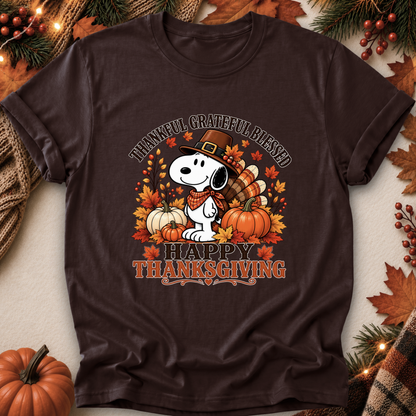 A dark brown short-sleeve graphic T-shirt laid flat with autumn props around it. The shirt features a Snoopy Thanksgiving design with the text 'THANKFUL GRATEFUL BLESSED' and 'HAPPY THANKSGIVING,' plus pumpkins, fall leaves, and a turkey.