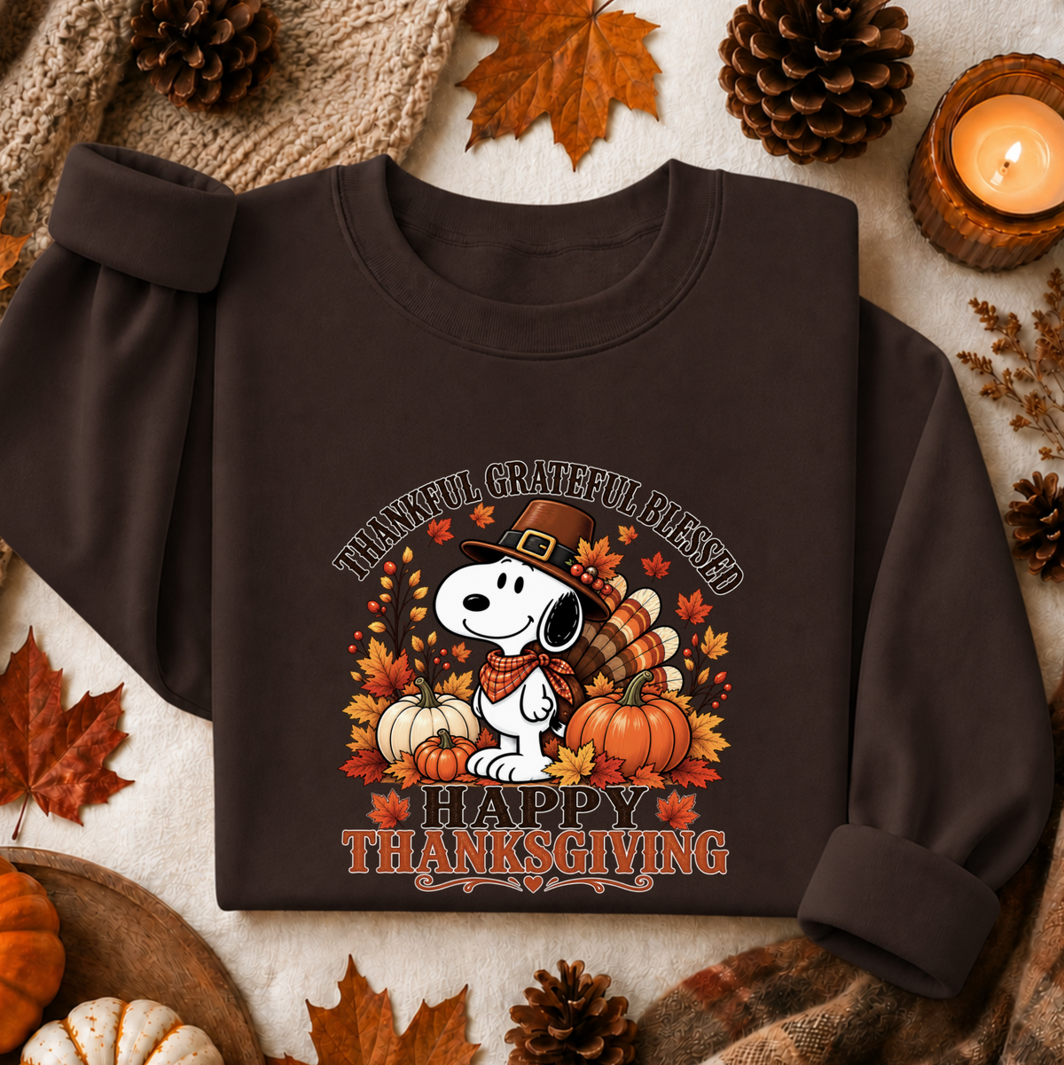 A dark brown crewneck sweatshirt featuring a festive Thanksgiving graphic of Snoopy with pumpkins, autumn leaves, and the text 'THANKFUL GRATEFUL BLESSED HAPPY THANKSGIVING'. The sweatshirt is styled in a flat-lay arrangement with pinecones, fall leaves, a candle, and a cozy knit blanket.