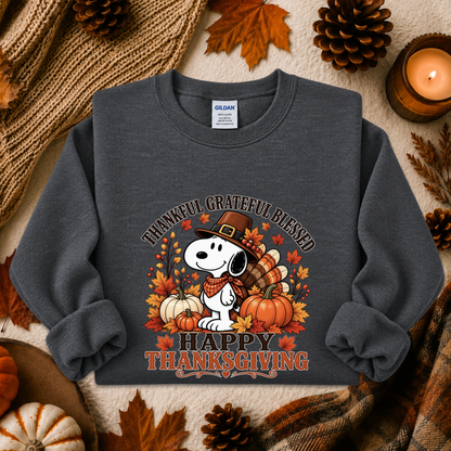 A dark gray crewneck sweatshirt featuring a Snoopy Thanksgiving graphic, styled in a flat lay with autumn props including pinecones, a candle, and a knitted blanket.