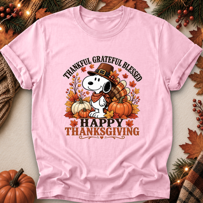 A light pink short-sleeve t-shirt with a Thanksgiving-themed graphic featuring Snoopy, pumpkins, autumn leaves, and the text 'THANKFUL GRATEFUL BLESSED HAPPY THANKSGIVING'. The shirt is displayed in a flat-lay arrangement with fall decorations including pine branches, berries, and a small pumpkin.