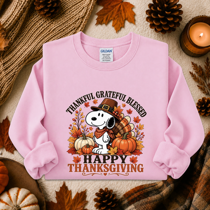 A light pink long-sleeve sweatshirt laid flat on a cozy autumn-themed background. The sweatshirt features a graphic of Snoopy wearing a pilgrim hat with pumpkins, fall leaves, and the text 'THANKFUL GRATEFUL BLESSED' and 'HAPPY THANKSGIVING.