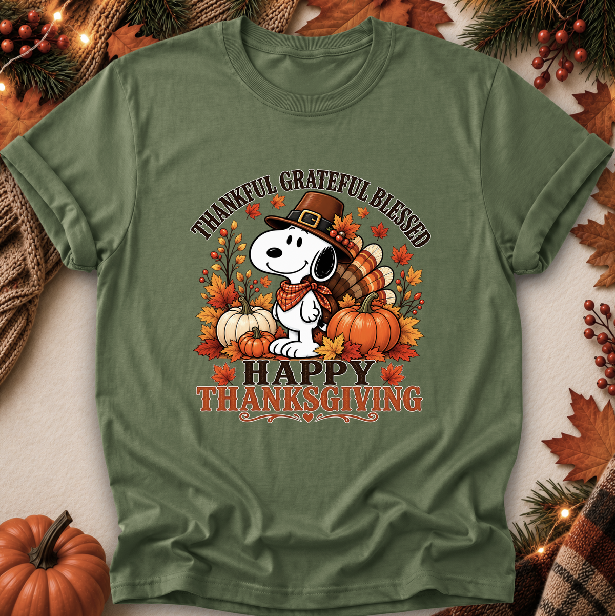 An olive green short-sleeve t-shirt featuring a Thanksgiving-themed graphic of Snoopy wearing a pilgrim hat with pumpkins, autumn leaves, and the text 'THANKFUL GRATEFUL BLESSED HAPPY THANKSGIVING'. The shirt is styled in a flat-lay with fall decorations including pine branches, berries, and a small pumpkin.